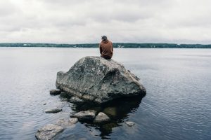 a man in a jacket sitting alone on a rock surrounded by water